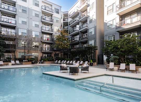 A swimming pool surrounded by lounge chairs in front of apartment buildings.