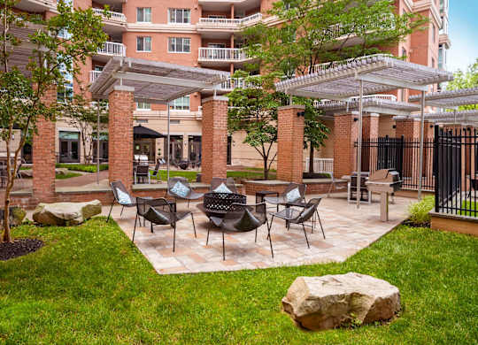 A patio with chairs and a table is surrounded by a grassy area and a building in the background.