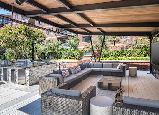 Covered outdoor lounge with gray sectional seating, white side tables, a television and nearby grilling stations.