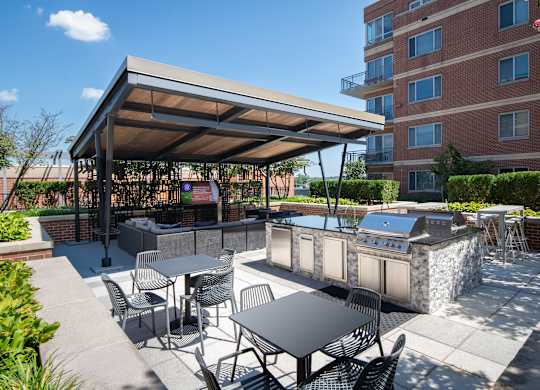 Covered outdoor kitchen with stainless steel grills, stone counters, bar seating, dining tables and a mounted television.