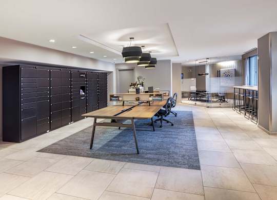 Vantage Mosaic lobby package area with black package lockers, a long wood table, chairs and light tile flooring.