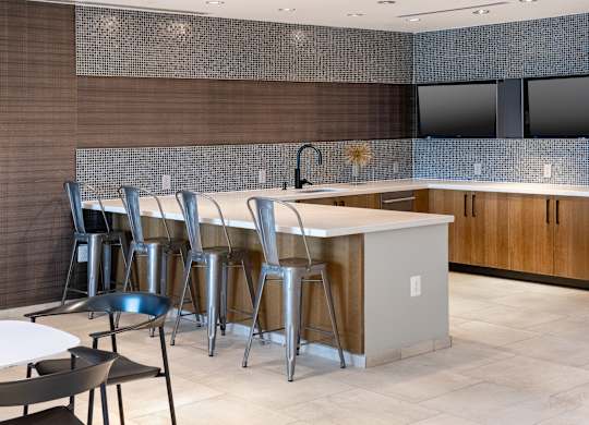 Vantage Mosaic lounge kitchen with wood cabinetry, patterned tile backsplash, white island and four metal barstools.