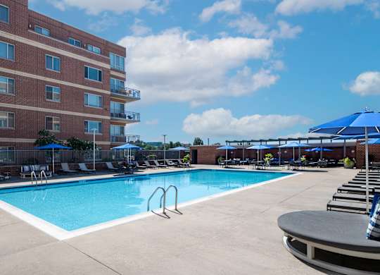 Vantage Mosaic swimming pool with metal handrails, rows of lounge chairs, blue umbrellas and a partly cloudy sky.