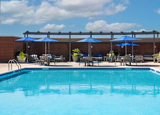 Low-angle view across the outdoor swimming pool toward blue umbrellas, tables, lounge chairs and the sundeck.