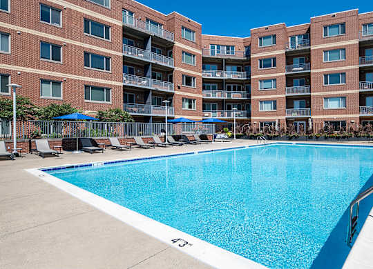 Large rectangular swimming pool at Vantage Mosaic surrounded by lounge chairs, blue umbrellas and brick apartment buildings.