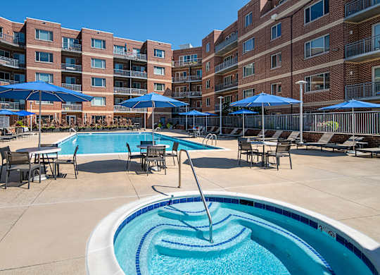 Round outdoor spa beside the Vantage Mosaic swimming pool, with tables, blue umbrellas and brick apartment buildings.