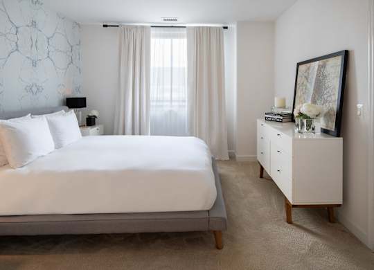 Bright model bedroom with a white bed, gray upholstered frame, white dresser, floor-length curtains and patterned accent wall.