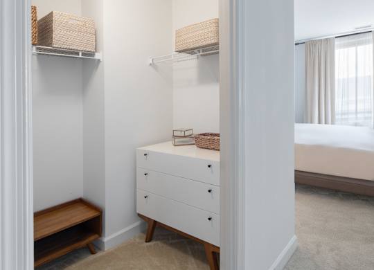 Open bedroom closet with white wire shelving, woven storage baskets, a small dresser and wood shoe rack.