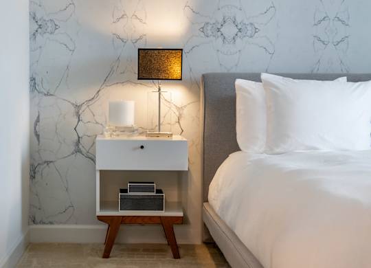 Close view of a model bedroom with a gray upholstered bed, white bedding, nightstand, black lamp and patterned wall.