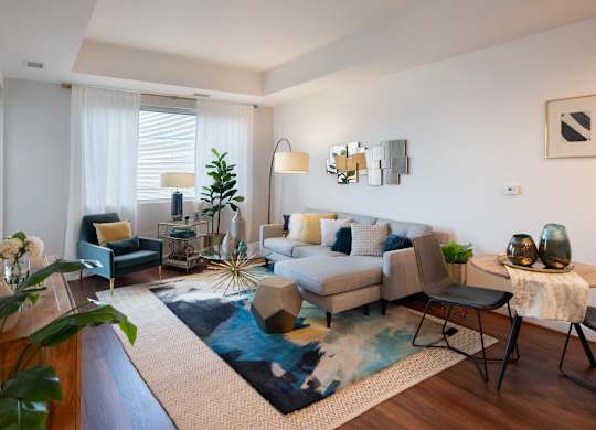 Sunlit model living room with a gray sectional, blue area rug, glass coffee table, accent chair and large windows.