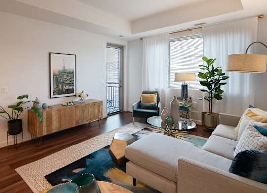 Model living room with a gray sectional, glass coffee table, wood media console, accent chair, plants and large windows.