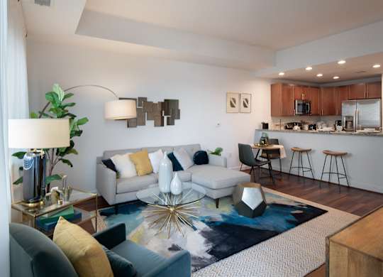 Open-concept model living room with a gray sectional, blue patterned rug, glass coffee table, breakfast bar and kitchen beyond.