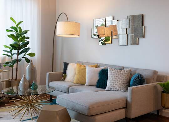 Close view of a gray sectional with colorful pillows, glass coffee table, arc floor lamp, wall art and indoor plants.