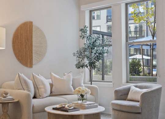 A living room with a white couch, a grey armchair, a round wooden table, and a large window with a view of the outside.