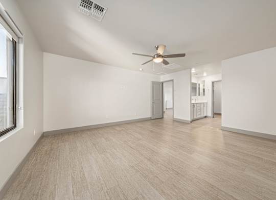 A spacious room with a ceiling fan and light, hardwood flooring, and a window on the left side.
