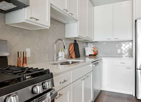 Hopkins, MN Luxury Apartments Near Methodist Hospital - The Hallon - Modern Kitchen with White Cabinetry, White Countertops, White Tile Backsplash, a Large Cooking Island, Hanging Lights, Stainless Steel Appliances, and Wood-Finish Plank Flooring