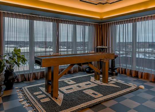 A large wooden table is in the middle of a room with a checkered rug.