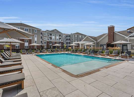 A large swimming pool surrounded by lounge chairs and umbrellas in front of apartment buildings.