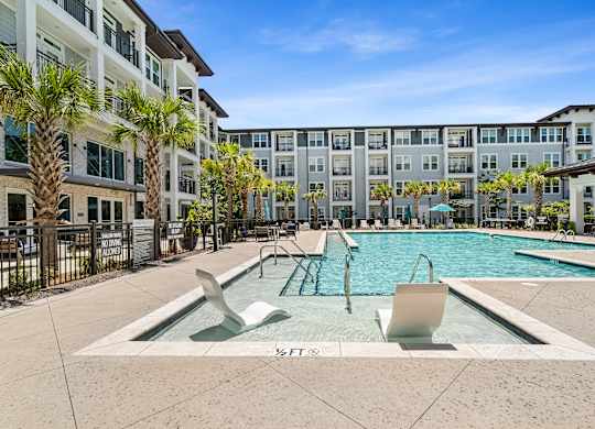 A swimming pool with two loungers in front of a building with palm trees.