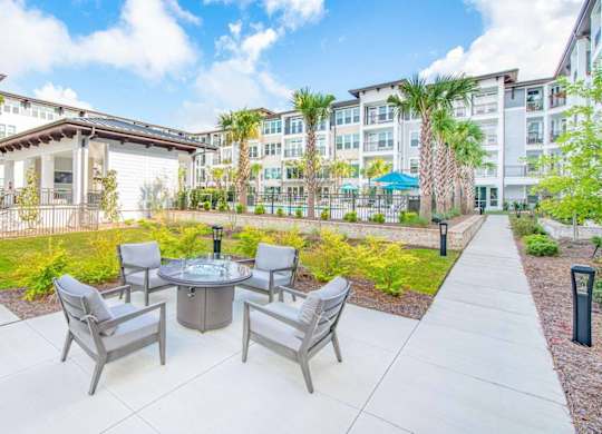 A patio with a table and chairs is surrounded by a grassy area and buildings.