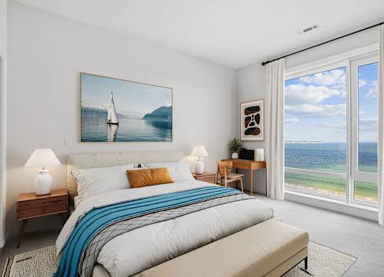 Carpeted penthouse bedroom with nightstands and a desk set beside a tall window over the sand and the open water.