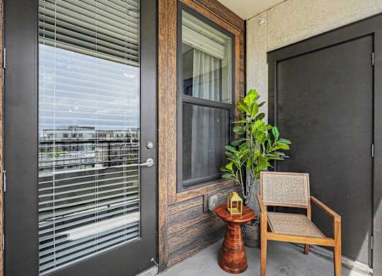 Balcony with a full glass door and window framed in wood-tone siding, a cane-back chair, side table, and a potted plant.