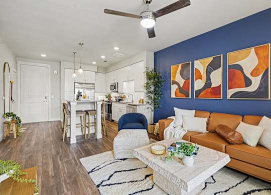Open living room with a ceiling fan and wood-look floors beside a white kitchen with pendants and an island with stools.