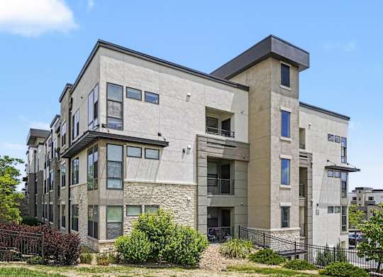 Apartment building with a stone base, stucco upper floors, stacked balconies, and landscaped grounds along a black fence.