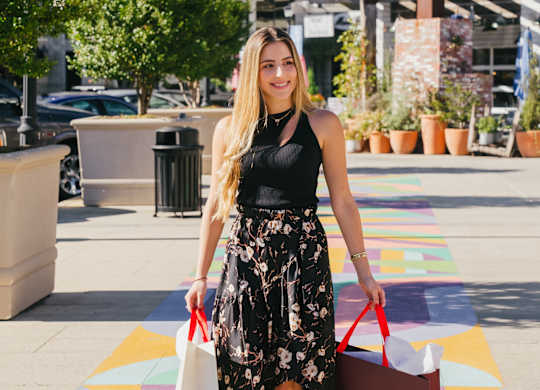 A woman in a black top and floral skirt is walking with shopping bags.