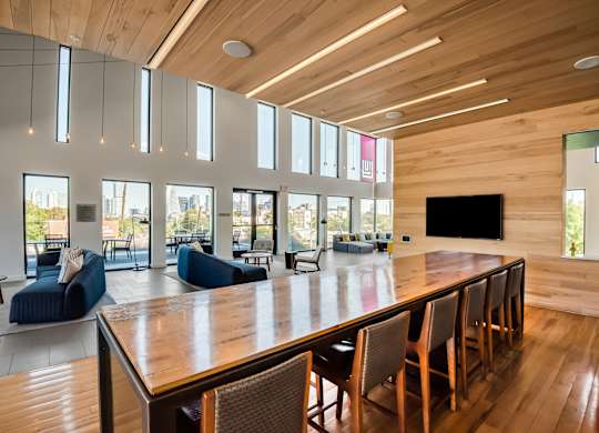 A long wooden table is surrounded by chairs in a room with a high ceiling and large windows.