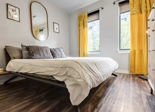 A bedroom with a bed, a mirror, and a window with yellow curtains.