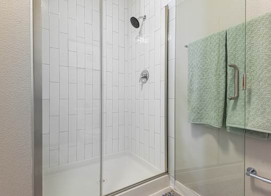 A white tiled shower with a glass door and a green towel hanging on the wall.