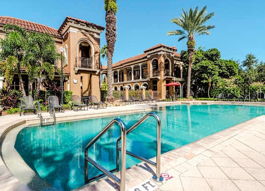Sparkling swimming pool framed by palm trees and Mediterranean-style buildings.