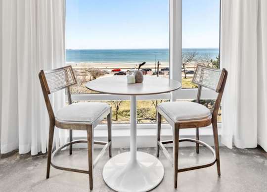 A dining table with chairs is set up by a window with a view of the beach.