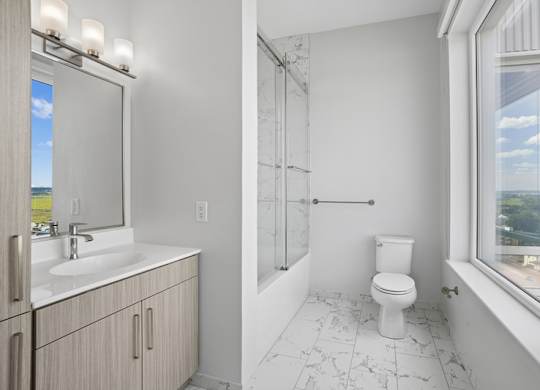 Penthouse bathroom with a glass-enclosed tub and shower, a quartz vanity, marble-look tile flooring and a window.