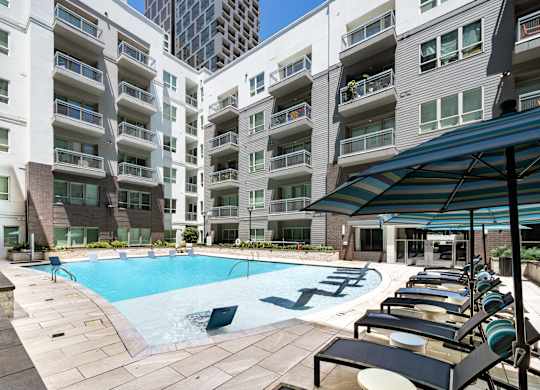 A swimming pool surrounded by lounge chairs and umbrellas in front of apartment buildings.