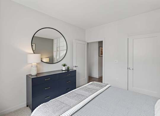 Apartment bedroom with a bed, dark dresser, round mirror, and two interior doors.