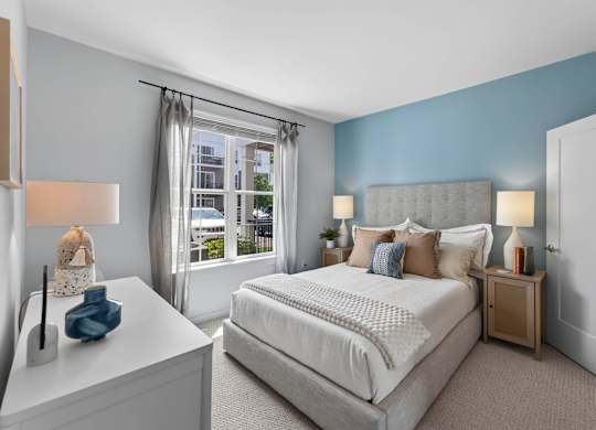 Apartment bedroom with a blue accent wall, upholstered bed, bedside tables, and window.