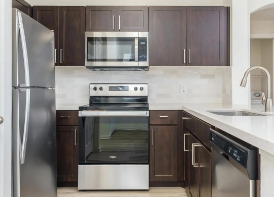 Apartments For Rent In Rancho Cucamonga, CA - Villas At The Gardens - Kitchen With Wood Plank Flooring, Stainless Steel Appliances, Dark Brown Wood-Tone Cabinets, Quartz Countertops, And Tile Backsplash.