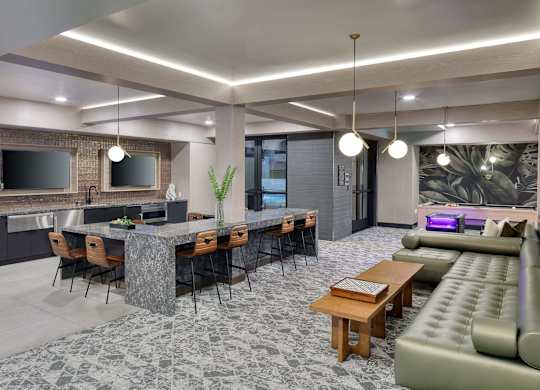 Game lounge with a long stone bar table, two mounted screens, a kitchenette and a leather sofa beyond at the far end.