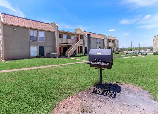 A barbecue grill is in the foreground of a grassy area in front of apartment buildings.