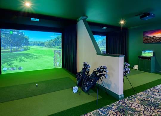 A golf simulator room with a screen, golf clubs, and a golf ball on the green carpet.