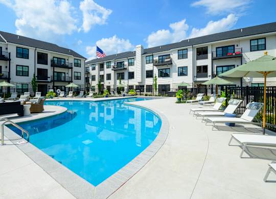 A swimming pool surrounded by lounge chairs and umbrellas in front of apartment buildings.