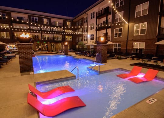 the pool is lit up at night with red chairs