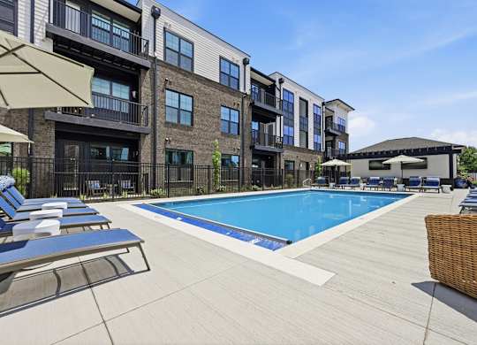 A large swimming pool is surrounded by sun loungers and umbrellas in front of a modern building.