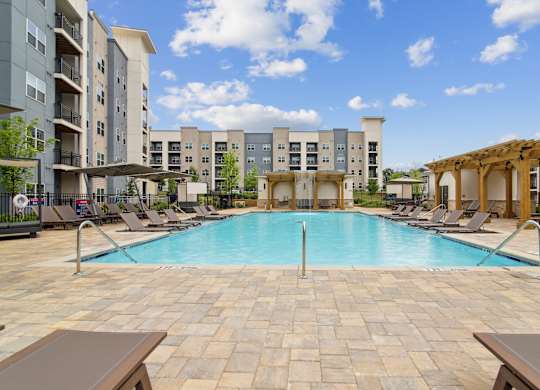 A large swimming pool surrounded by lounge chairs and umbrellas in front of apartment buildings.