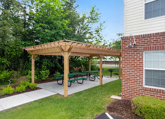 A wooden pergola with green benches is in front of a brick building.