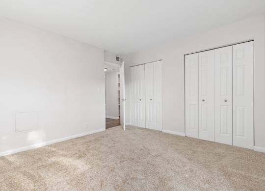 A room with white walls and carpeted floor with a doorway leading to another room.