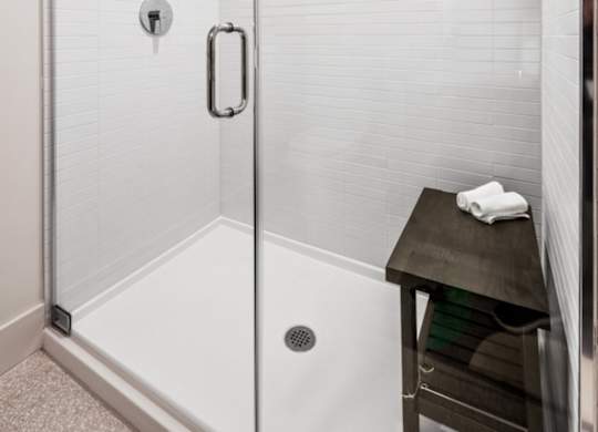 A white tiled shower with a glass door and a black stool.