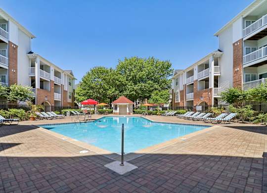 A swimming pool surrounded by a brick patio and lounge chairs with a red umbrella in the middle.
